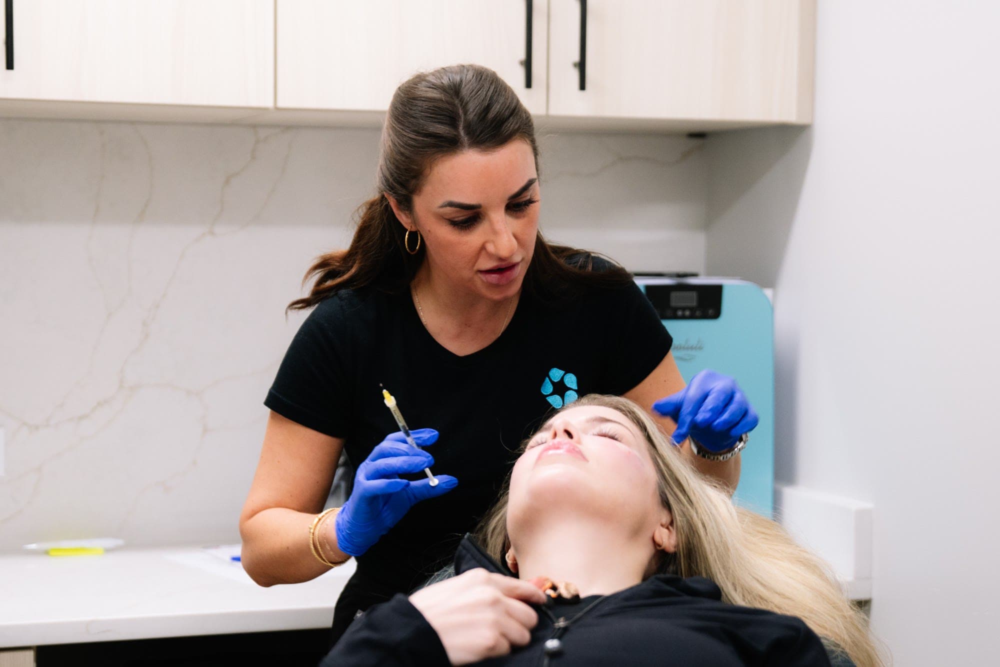 An injector explains the plan to a reclining patient before jawline treatment